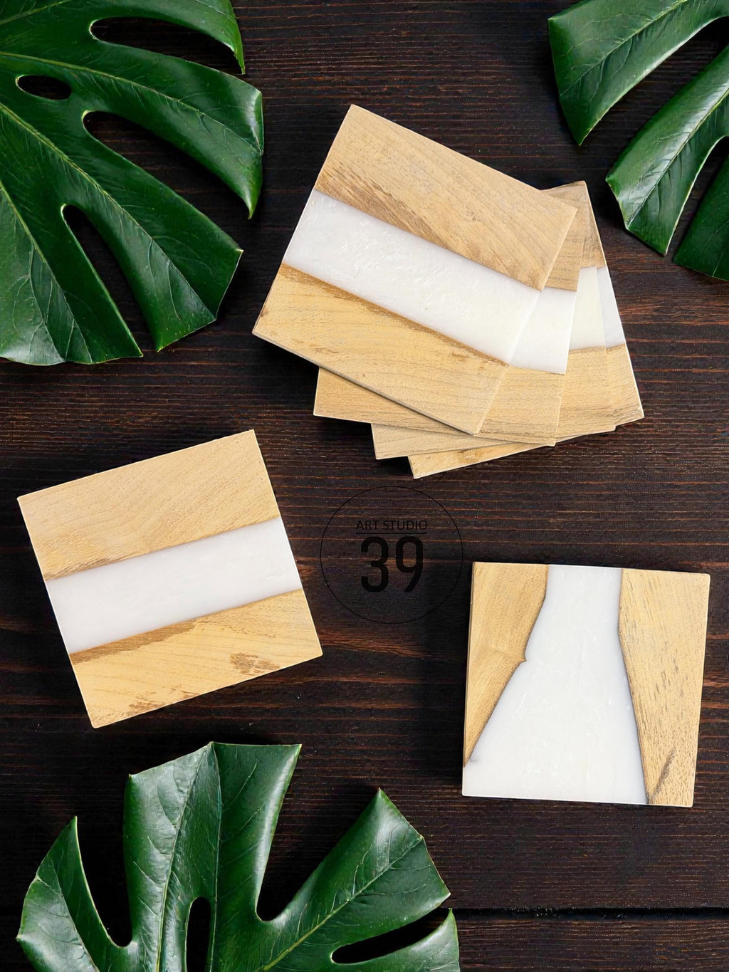 Wooden square coasters with a light wood grain and a white resin inlay. The coasters are arranged on a dark wood surface, with green leaves in the background. The coasters have a modern, minimalist design.