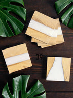 Wooden square coasters with a light wood grain and a white resin inlay. The coasters are arranged on a dark wood surface, with green leaves in the background. The coasters have a modern, minimalist design.