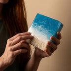 A rectangular art piece depicting a beach scene. The top portion is a translucent blue, resembling the ocean, with white swirls. The bottom section is beige, mimicking sand. The piece is held in hands.