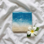 Square resin art piece depicting a beach scene with blue ocean and white waves over a sandy shore. A white flower with yellow center and green leaves sits next to the art. The text "ART STUDIO 39" is in a circle.