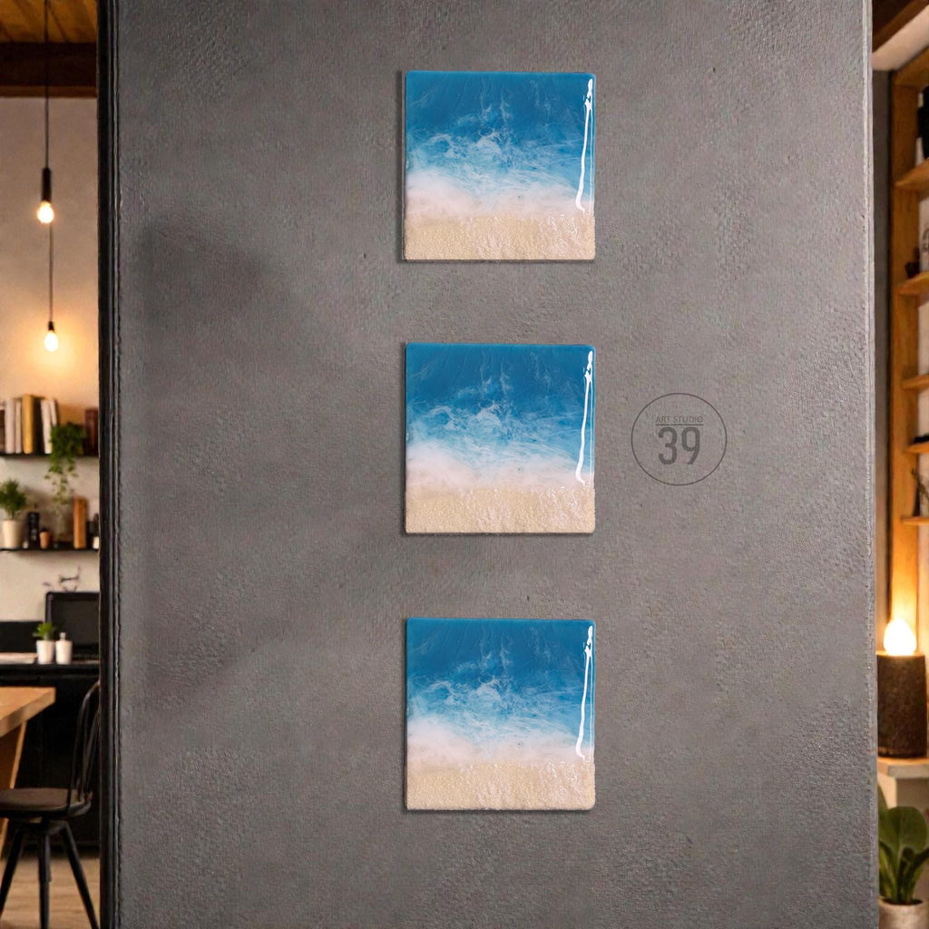 Three square wall art pieces depicting a beach scene with blue ocean, white waves, and a sandy shore. The artwork is mounted on a gray wall. The art pieces are arranged vertically.