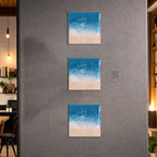 Three square wall art pieces depicting a beach scene with blue ocean, white waves, and a sandy shore. The artwork is mounted on a gray wall. The art pieces are arranged vertically.