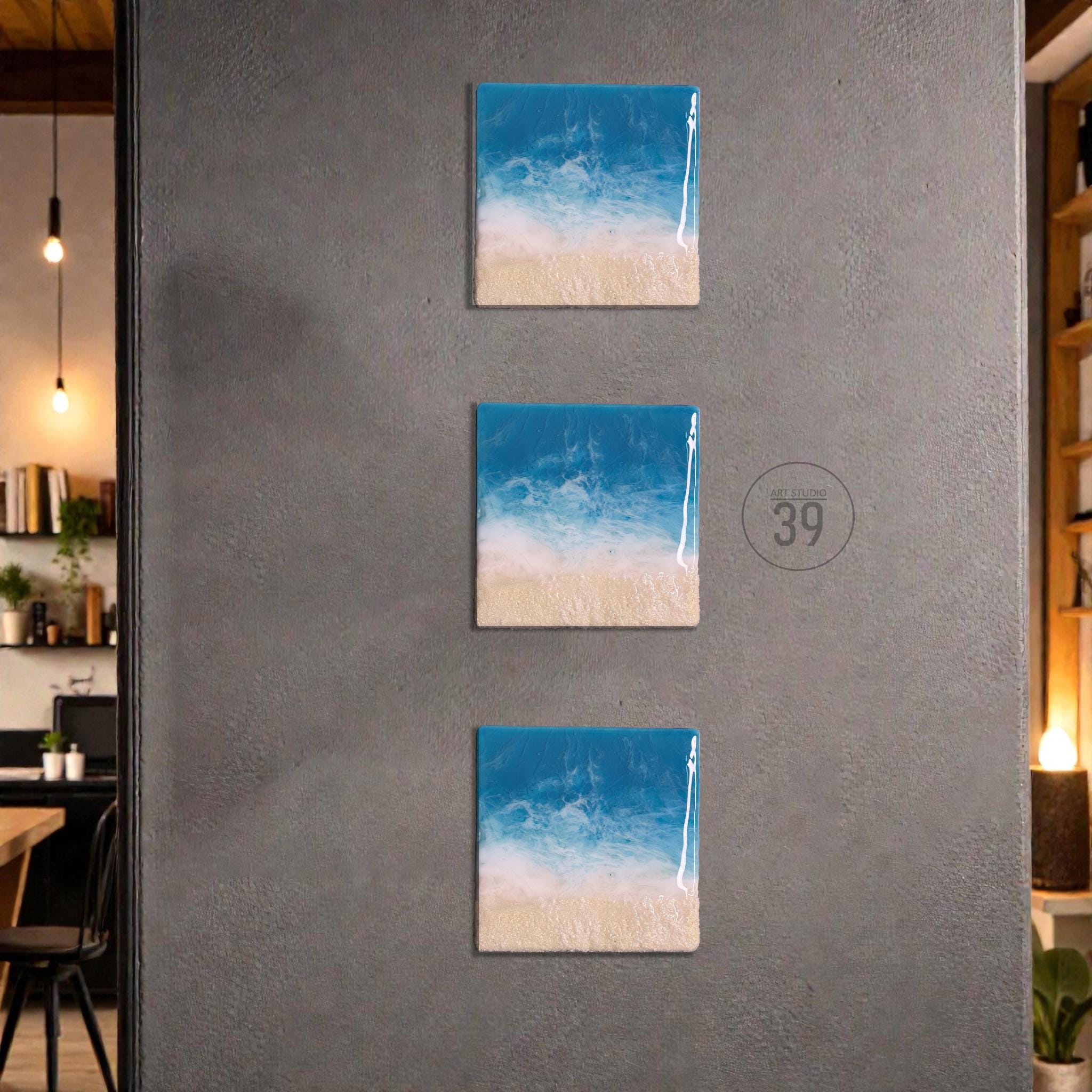 Three square wall art pieces depicting a beach scene with blue ocean, white waves, and a sandy shore. The artwork is mounted on a gray wall. The art pieces are arranged vertically.