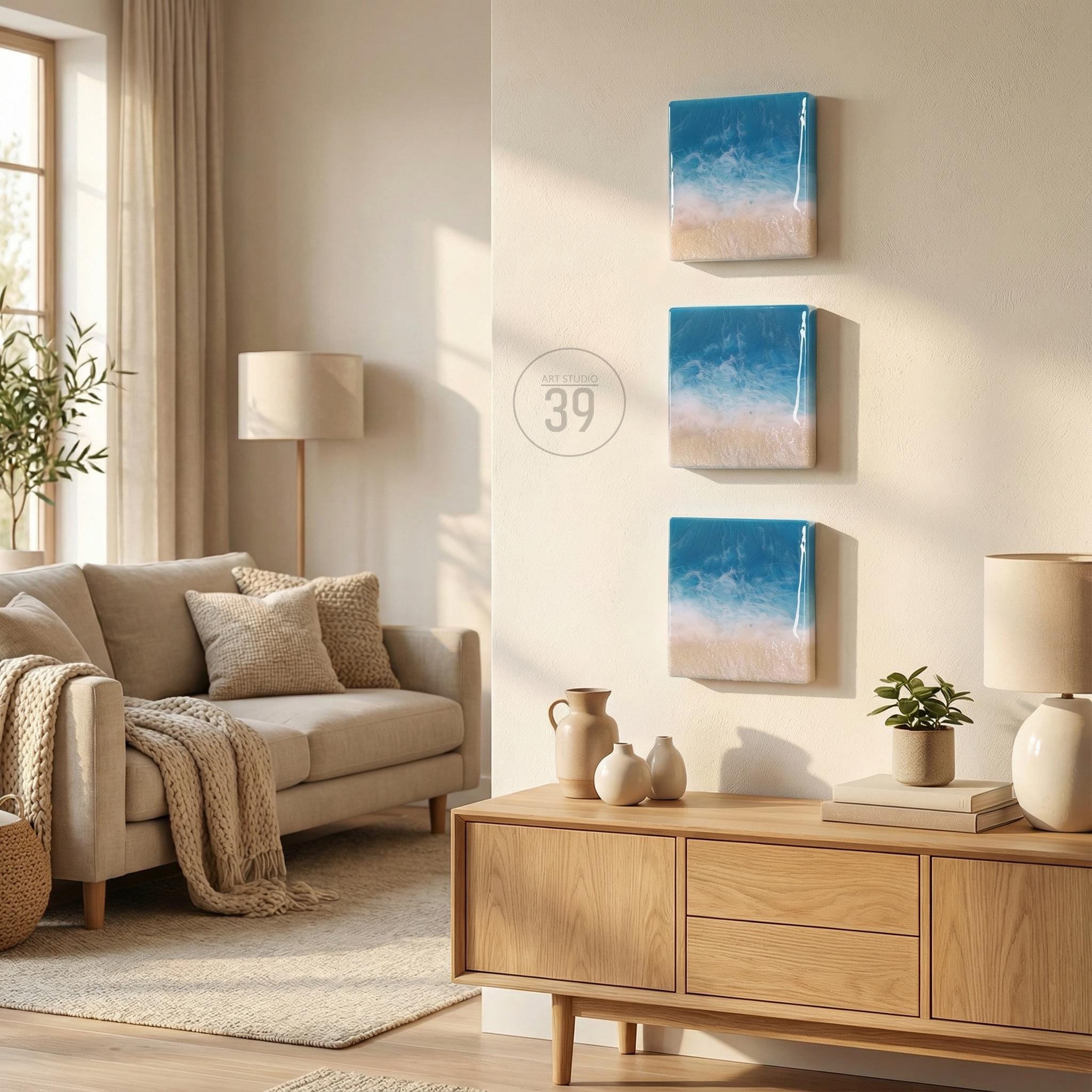 Three square wall art pieces depicting a beach scene with blue, white, and tan colors. The art is mounted on a light-colored wall. The room also features a beige sofa, wooden cabinet, and decorative items.