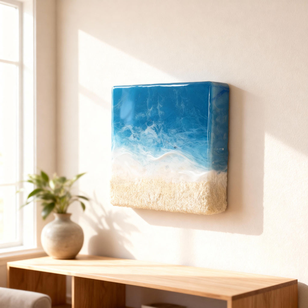 Square wall art depicting a beach scene. The top portion is a vibrant blue, transitioning to white, resembling ocean waves. The bottom section is textured, sandy beige. The artwork is mounted on a neutral-colored wall.