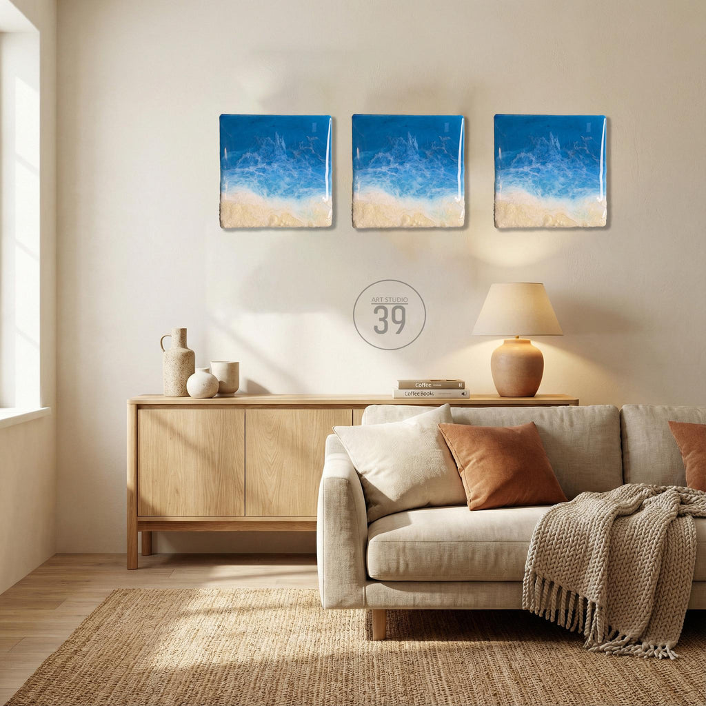 Three square wall art pieces depicting a beach scene with blue ocean and sandy shore. The art has a textured appearance. The text "ART STUDIO 39" is in a circle below the art. The room has a neutral color palette with a sofa, rug, and wooden cabinet.