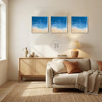 Three square wall art pieces depicting a beach scene with blue ocean and sandy shore. The art has a textured appearance. The text "ART STUDIO 39" is in a circle below the art. The room has a neutral color palette with a sofa, rug, and wooden cabinet.
