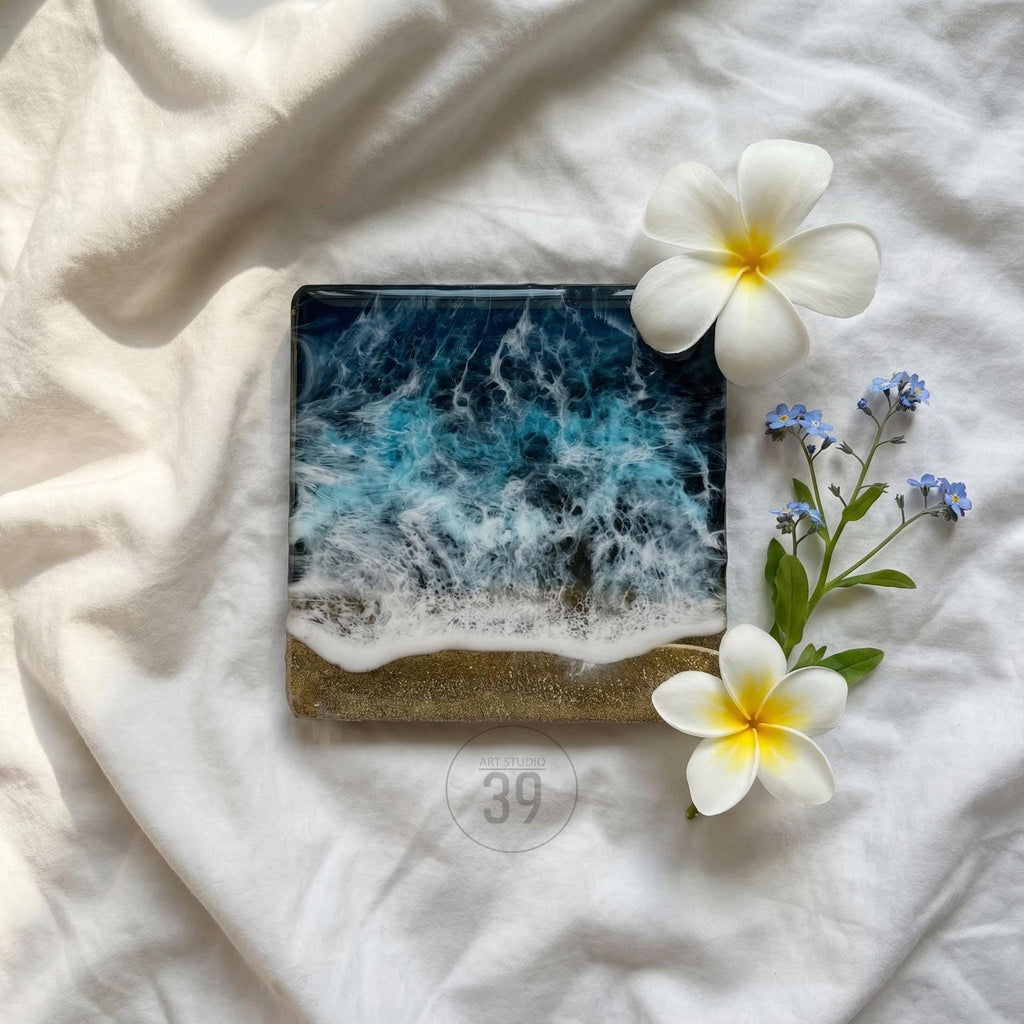 Square resin art piece depicting an ocean scene with blue and white waves crashing onto a sandy shore. The artwork is adorned with white plumeria flowers and small blue forget-me-nots. The piece has the text "39" in a circle.