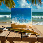 Square resin art piece depicting an ocean scene with blue and white waves crashing on a sandy beach. The artwork is displayed on a wooden table with a palm leaf and seashells. The background shows a beach and ocean.