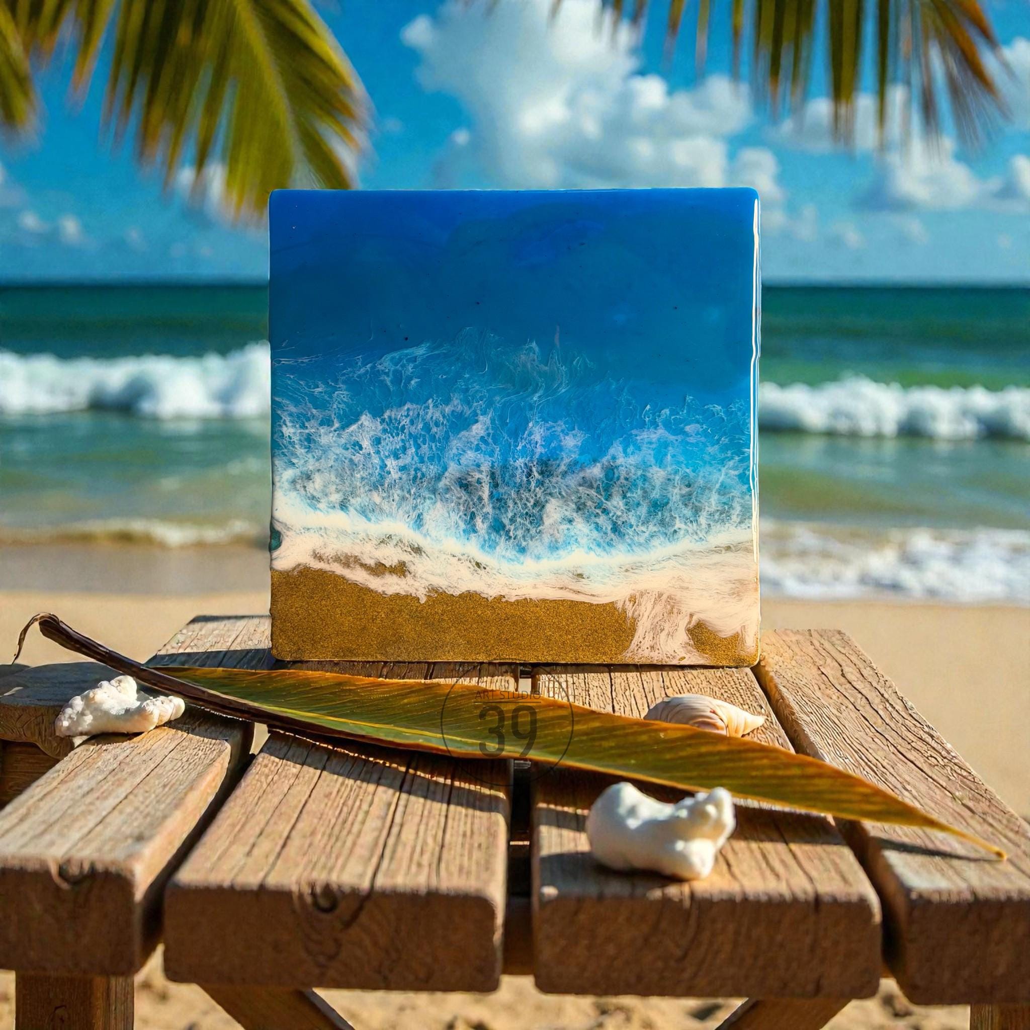 Square resin art piece depicting an ocean scene with blue and white waves crashing on a sandy beach. The artwork is displayed on a wooden table with a palm leaf and seashells. The background shows a beach and ocean.