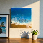 Square resin art piece depicting a beach scene with blue ocean waves, white foam, and golden sand. The artwork is mounted on a wall. A framed picture and a potted succulent are also visible.