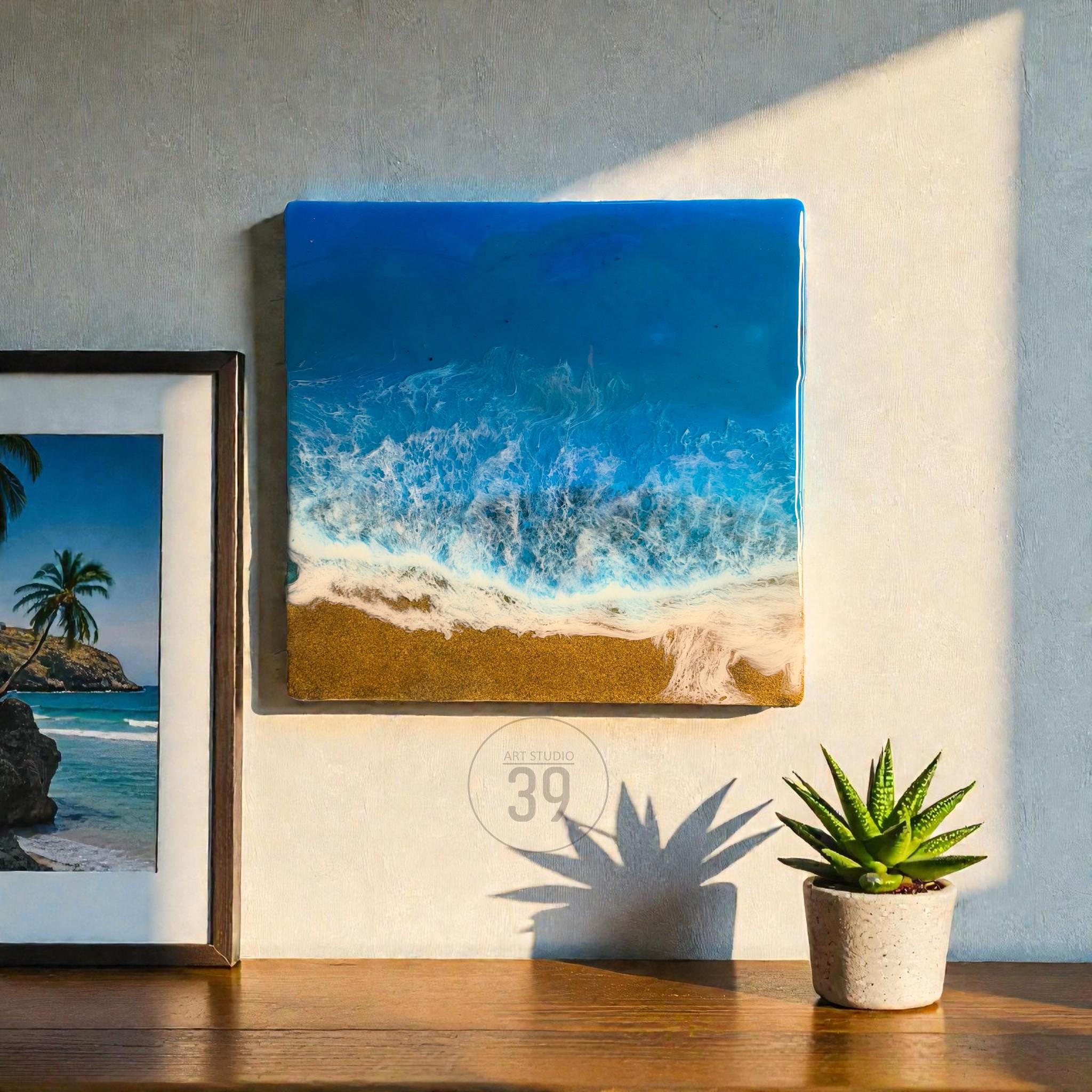 Square resin art piece depicting a beach scene with blue ocean waves, white foam, and golden sand. The artwork is mounted on a wall. A framed picture and a potted succulent are also visible.