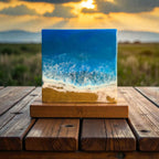 A square resin art piece depicting an ocean scene with blue waves, white foam, and a sandy beach. The artwork is mounted on a wooden base, with the number "39" visible. The background features a sunset over a field.