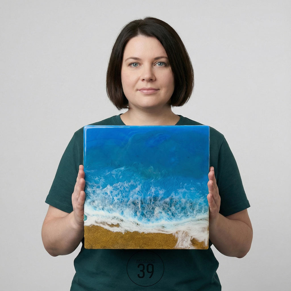 Square resin art piece depicting an ocean scene with blue and white waves crashing onto a sandy shore. The artwork has a glossy finish and is held by a person. The bottom right corner of the art piece has the number "39".