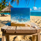 Square resin art piece depicting a beach scene with blue ocean waves and golden sand. The artwork is displayed on a wooden stool with seashells scattered around. The background shows a beach with palm trees and a straw umbrella.