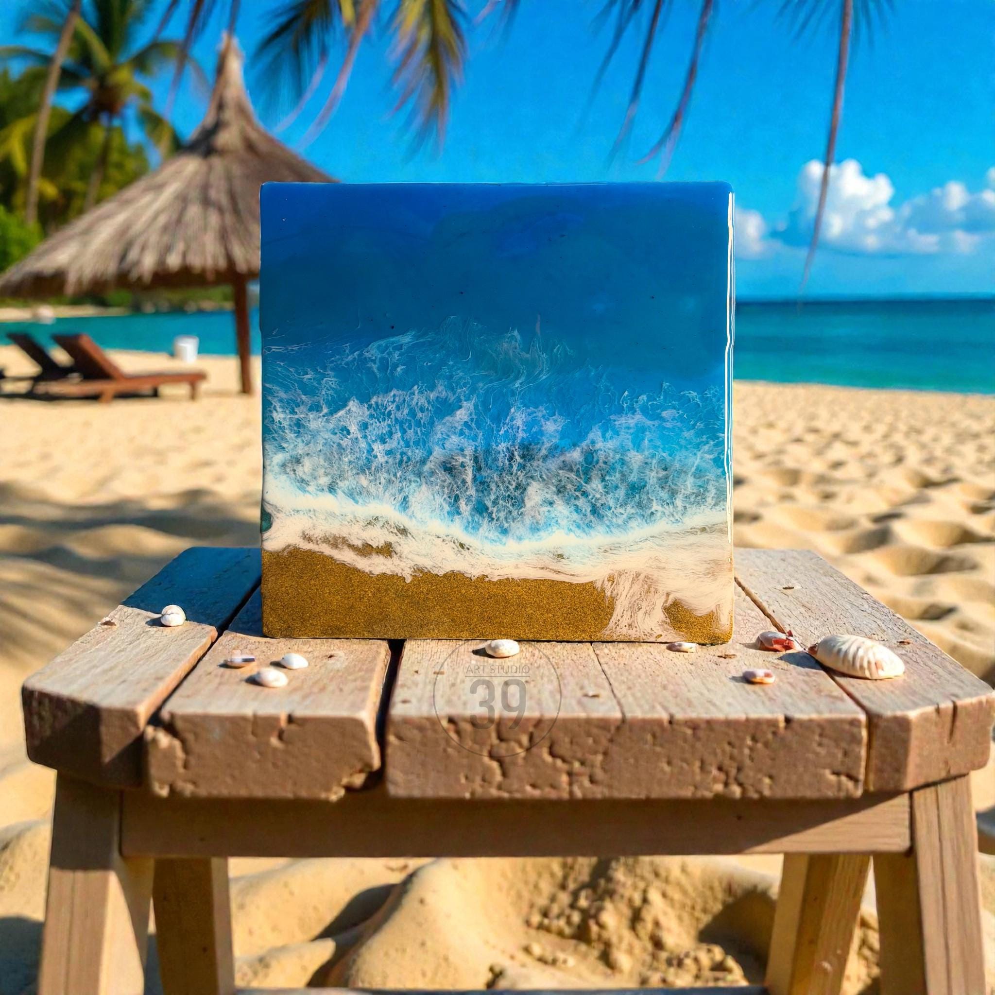 Square resin art piece depicting a beach scene with blue ocean waves and golden sand. The artwork is displayed on a wooden stool with seashells scattered around. The background shows a beach with palm trees and a straw umbrella.