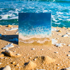 Square resin art piece depicting an ocean scene. The artwork features layers of blue and white resin to mimic waves, with a sandy beach at the bottom. The piece is set on a sandy beach with ocean waves in the background.