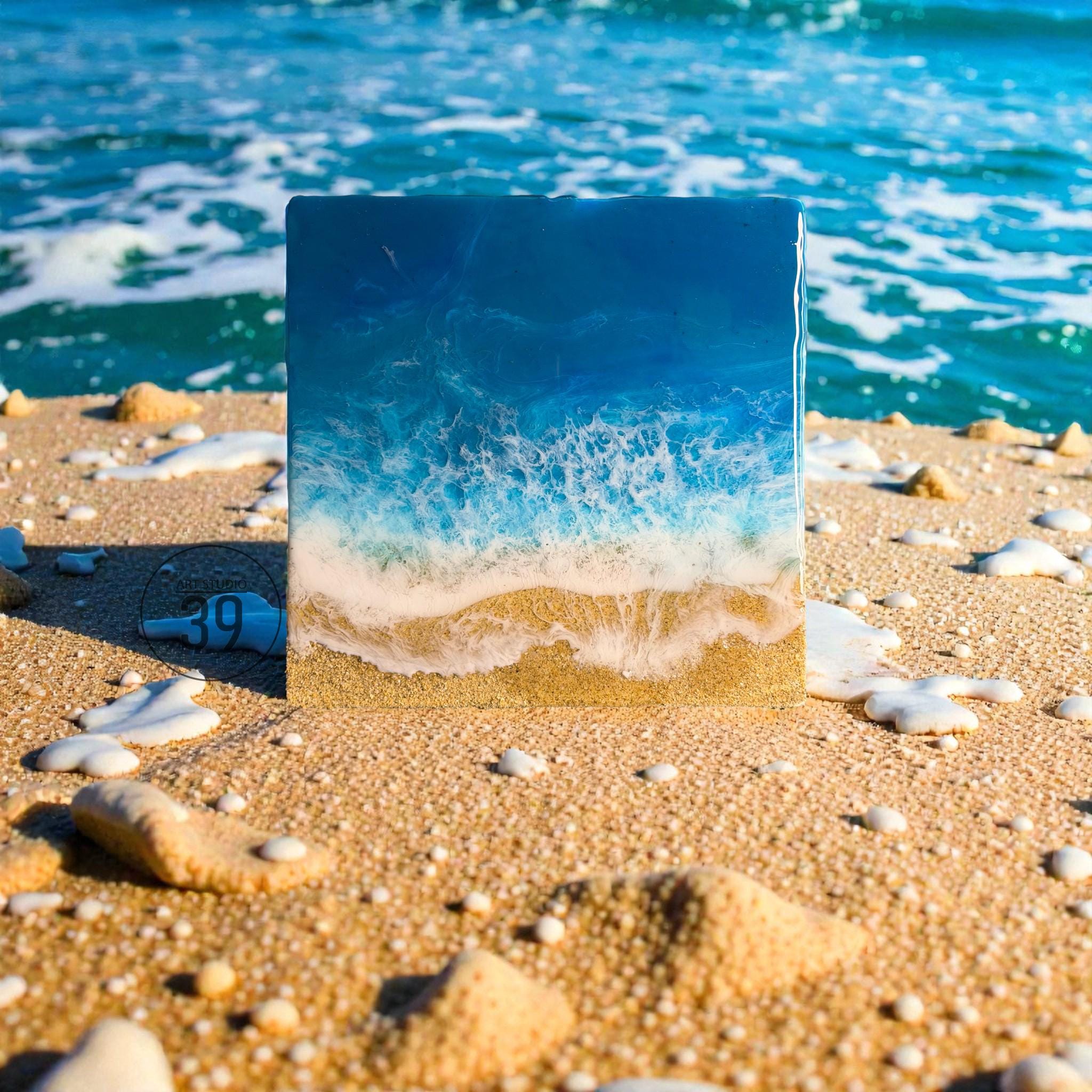 Square resin art piece depicting an ocean scene. The artwork features layers of blue and white resin to mimic waves, with a sandy beach at the bottom. The piece is set on a sandy beach with ocean waves in the background.