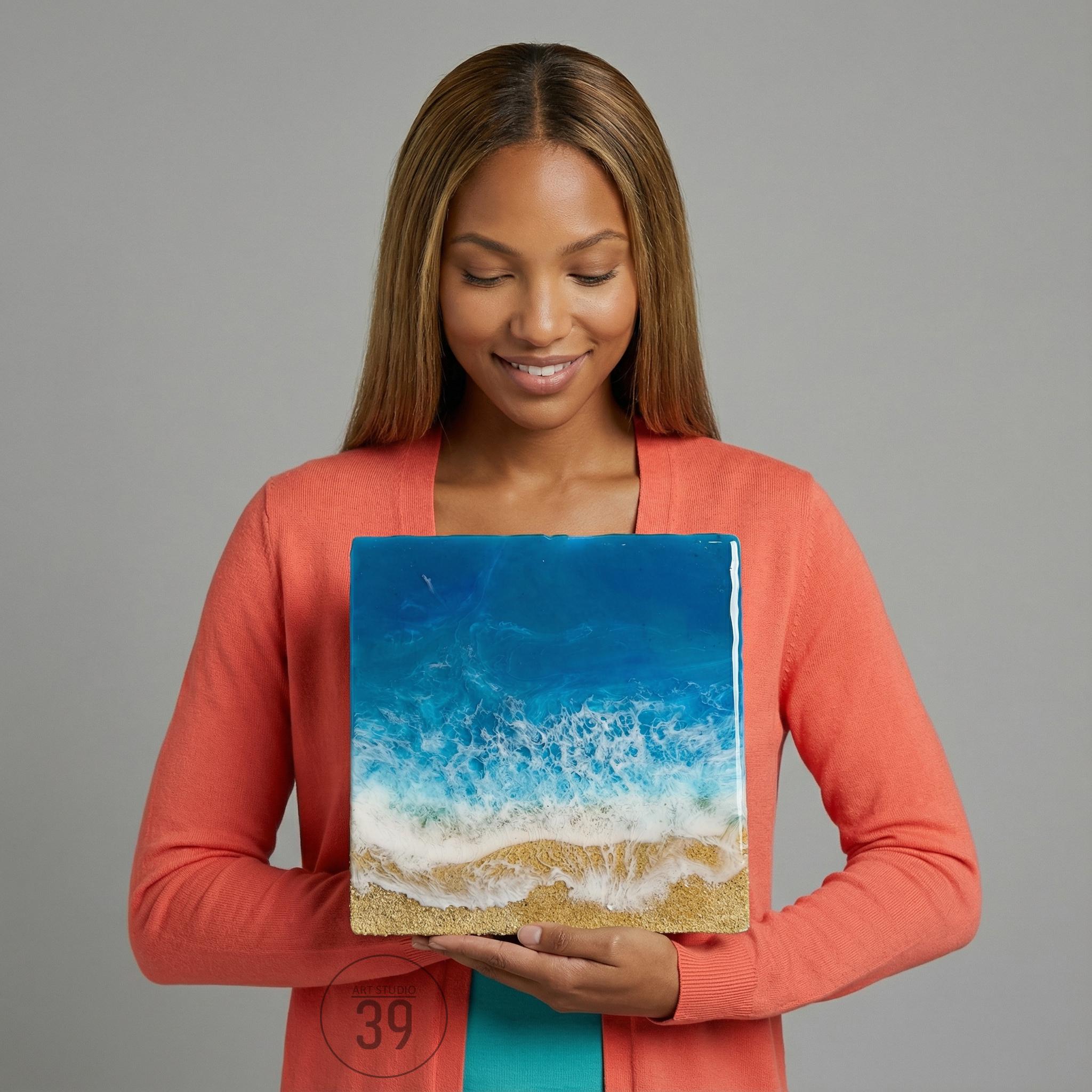 A square resin art piece depicting an ocean scene with blue and white waves crashing onto a sandy beach. The artwork is held by a person wearing a coral-colored cardigan.