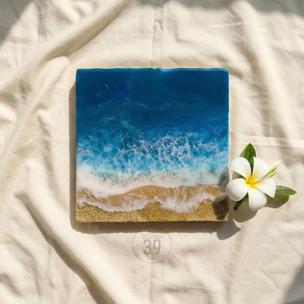 Square resin art piece depicting a beach scene. The top half is deep blue, transitioning to foamy white waves and golden sand at the bottom. A white flower with a yellow center and green leaves rests beside the art.