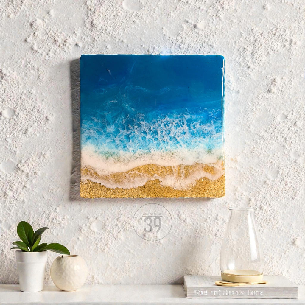 Square resin art piece depicting an ocean scene. The artwork features a gradient of blue hues representing the sea, transitioning to white foamy waves and a golden sand beach. The piece is mounted on a white textured wall.
