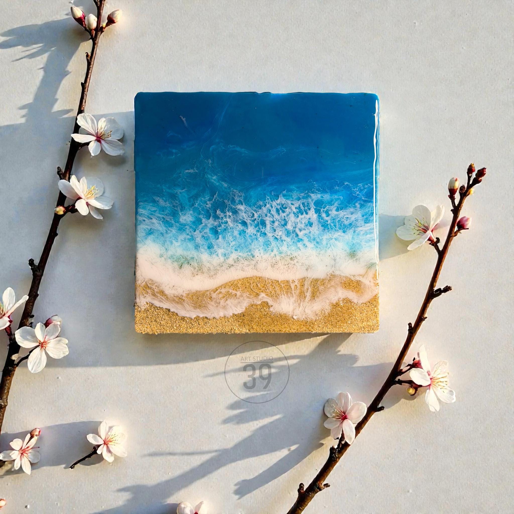 Square resin art piece depicting a beach scene. The artwork features a gradient of deep blue to white, representing the ocean, and a sandy beach at the bottom. White floral branches frame the artwork.