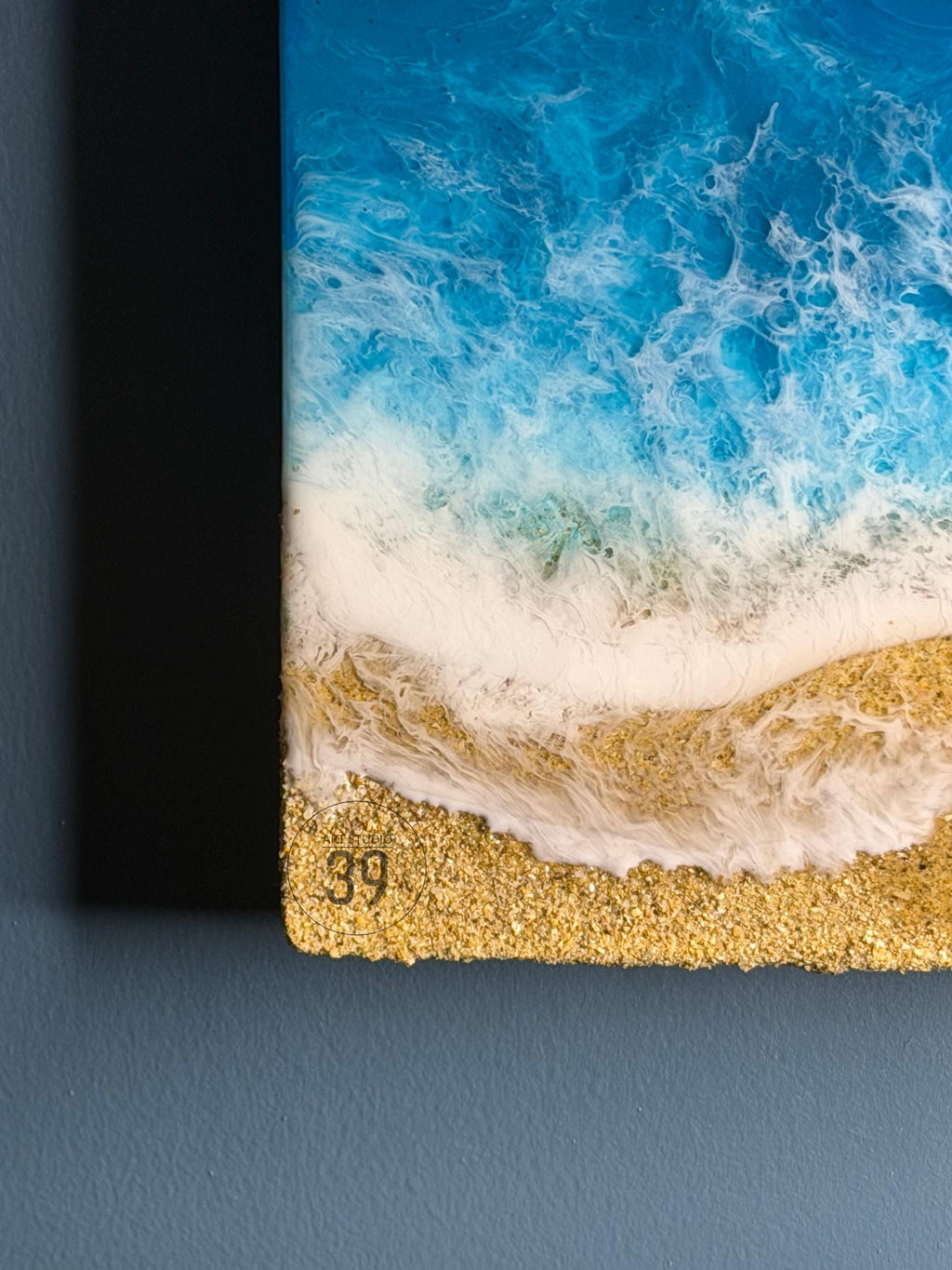 Abstract art piece depicting a beach scene. The artwork features a vibrant blue ocean with white foamy waves crashing onto a golden sand shore. The number "39" is visible in the bottom corner. The piece is likely made with resin.