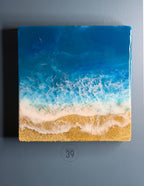 Square resin art piece depicting a beach scene. The top portion is a deep blue, transitioning to lighter blues and white, mimicking ocean waves. The bottom features golden sand. The art piece has the text "art studio 39".