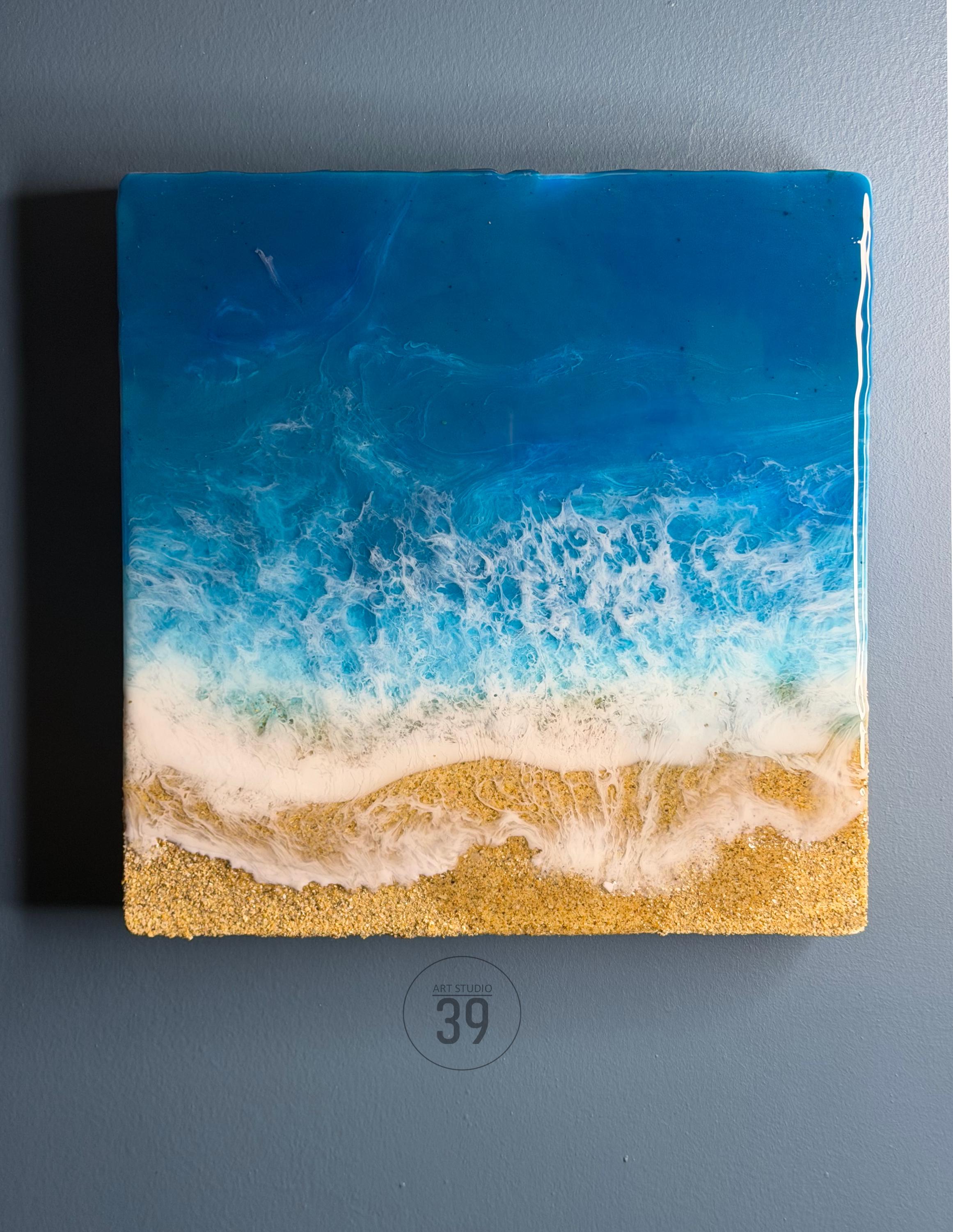 Square resin art piece depicting a beach scene. The top portion is a deep blue, transitioning to lighter blues and white, mimicking ocean waves. The bottom features golden sand. The art piece has the text "art studio 39".