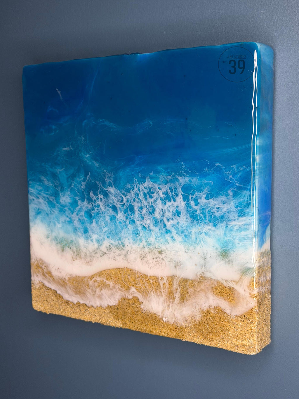 Square resin art piece depicting a beach scene. The artwork features a gradient of blues representing the ocean, white foamy waves, and a sandy beach. The number "39" is visible in the top right corner. The piece is mounted on a wooden frame.