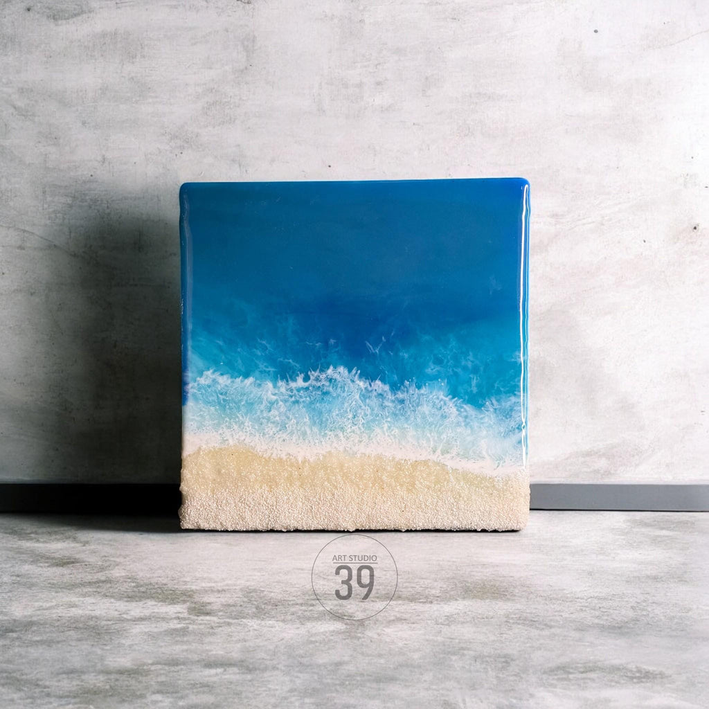 Square resin art piece depicting a beach scene. The artwork features a gradient of blues representing the ocean, transitioning to white foamy waves and a sandy beige beach. The text "ART STUDIO 39" is visible.