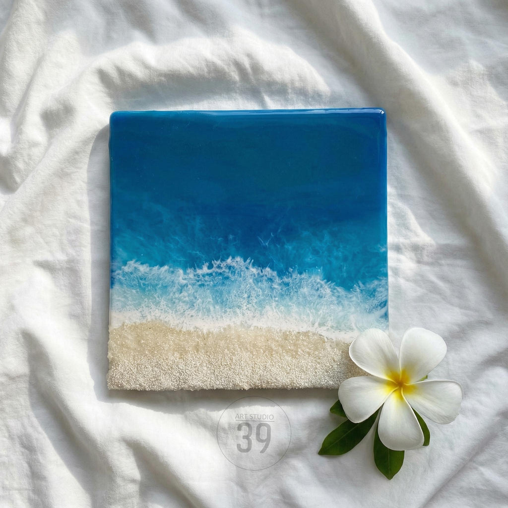 Square resin art piece depicting a beach scene with a gradient of blue ocean and white foamy waves meeting a sandy shore. A white flower with yellow center and green leaves are in the lower right corner.