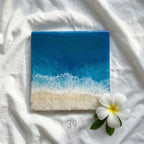 Square resin art piece depicting a beach scene with a gradient of blue ocean and white foamy waves meeting a sandy shore. A white flower with yellow center and green leaves are in the lower right corner.