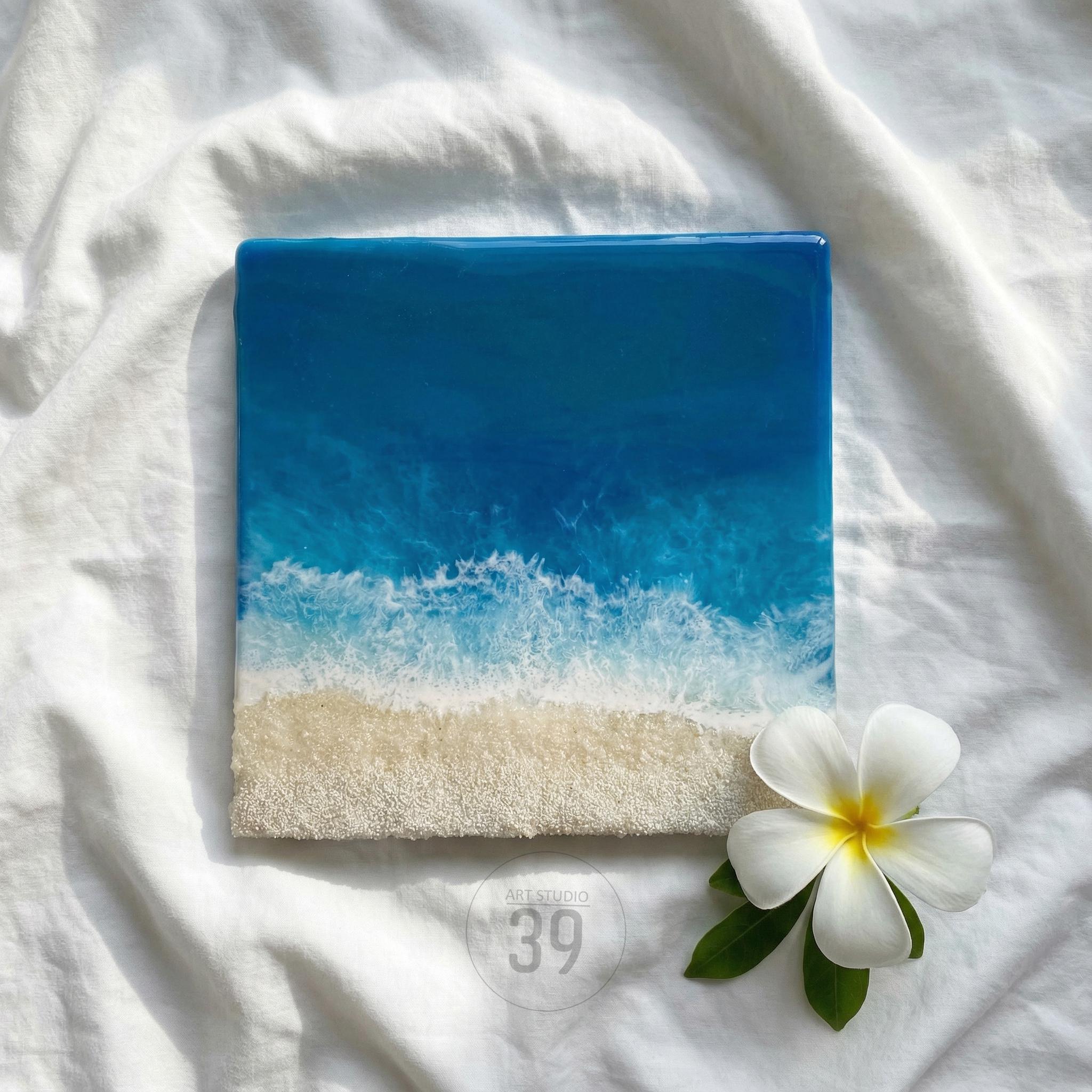 Square resin art piece depicting a beach scene with a gradient of blue ocean and white foamy waves meeting a sandy shore. A white flower with yellow center and green leaves are in the lower right corner.