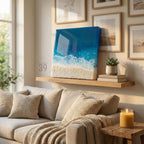 A square canvas art piece depicting a beach scene with blue ocean waves and white foam, transitioning to a sandy shore. The artwork is displayed on a wooden shelf above a beige sofa, with the text "39" visible.