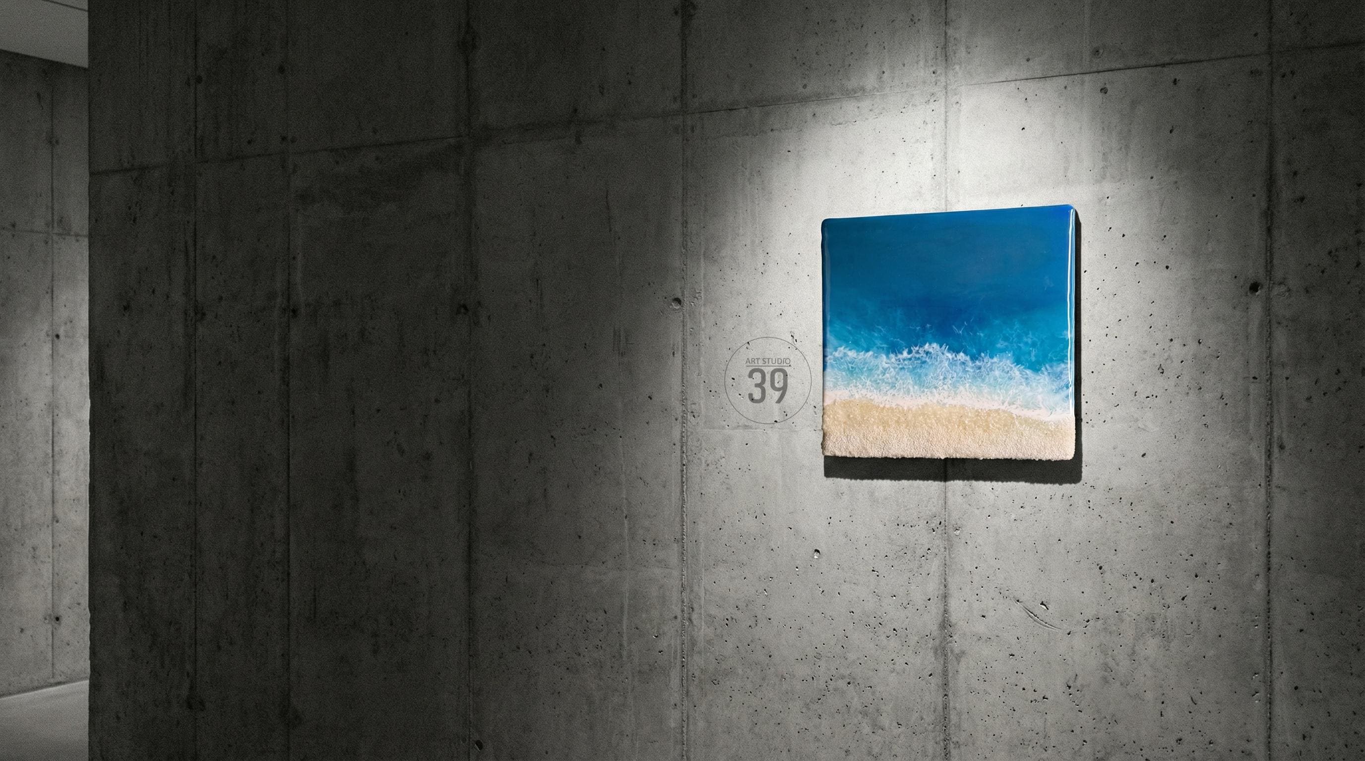 Square canvas art depicting a beach scene with a blue ocean, white waves, and a sandy shore. The artwork is mounted on a concrete wall. The number "39" is visible on the wall.