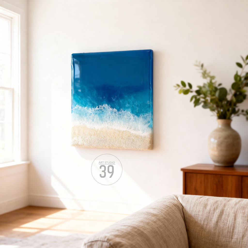 A square resin art piece depicting a beach scene. The artwork features a gradient of deep blue to white, representing the ocean and foamy waves, transitioning to a sandy beige beach. The piece is mounted on a white wall.