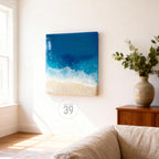 A square resin art piece depicting a beach scene. The artwork features a gradient of deep blue to white, representing the ocean and foamy waves, transitioning to a sandy beige beach. The piece is mounted on a white wall.
