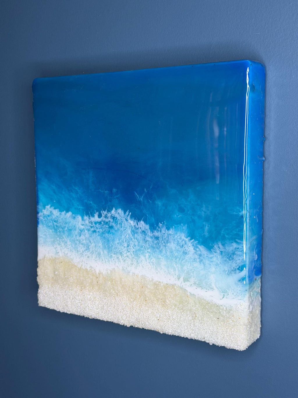 A square resin art piece depicting a beach scene. The top portion is a gradient of blues representing the ocean, transitioning to white waves and a sandy beach at the bottom. The artwork is mounted on a square canvas.