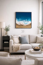 Square framed artwork depicting an aerial view of a beach scene. The painting features a deep blue ocean, white foamy waves, and a sandy shore. The artwork hangs above a light-colored sofa with decorative pillows.