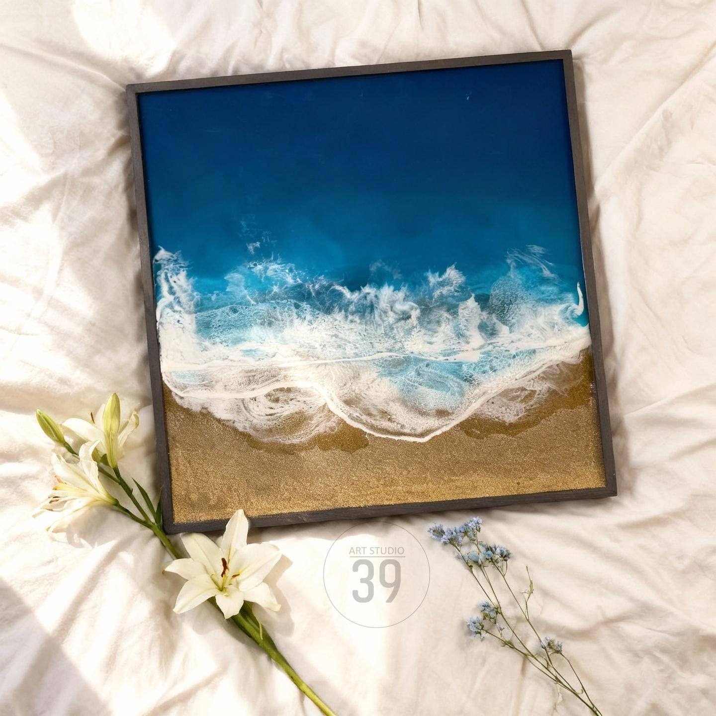 Square framed artwork depicting a beach scene with a dark blue ocean, white foamy waves, and a sandy shore. The artwork is framed in a dark gray frame. The text "ART STUDIO 39" is in a circle in the bottom right corner.