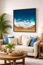 A framed artwork depicting a beach scene with a dark blue ocean, white foamy waves, and golden sand. The artwork hangs above a beige sofa with blue and striped pillows. A round wooden coffee table with a woven tray and decorative items sits in front of the sofa.