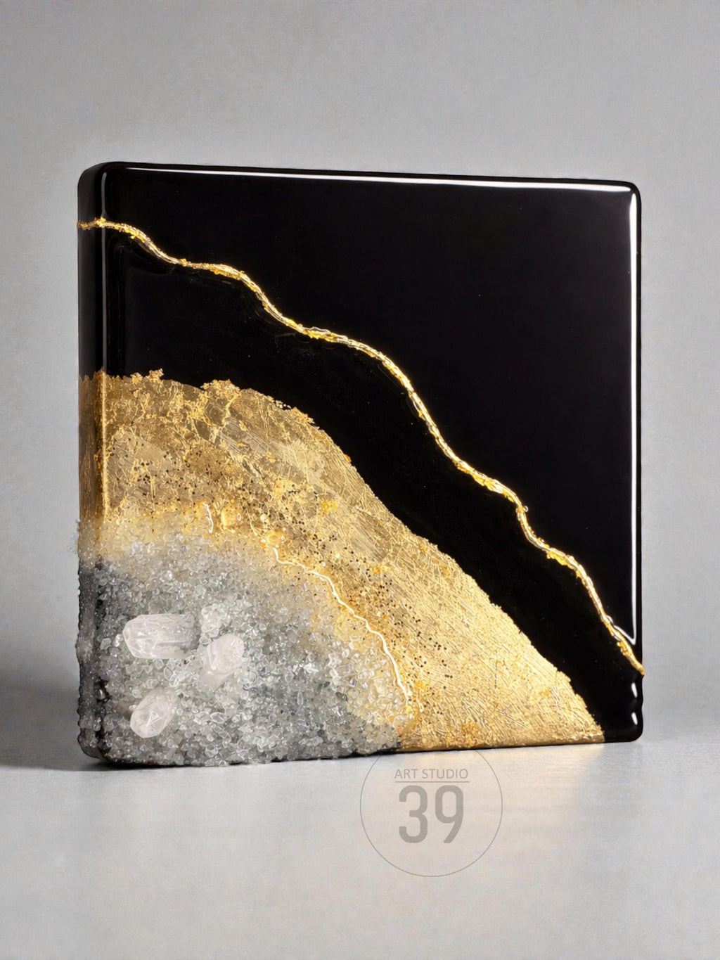 A square, abstract art piece with a glossy black top half and a textured bottom half. The bottom half transitions from gold to gray with embedded crystals. A gold line runs diagonally across the piece. The text "ART STUDIO 39" is in the bottom right corner.