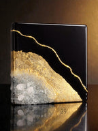 A square, black decorative object with a gold-colored, vein-like design. The bottom half features a textured, light gray and gold surface with embedded clear crystals. The object has a glossy finish.