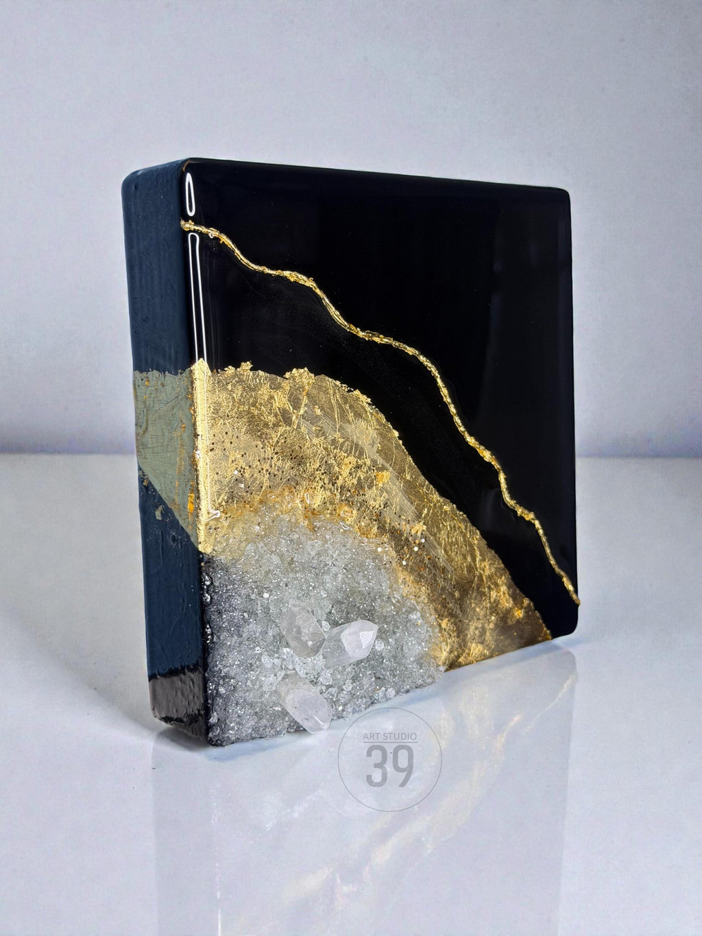 A square, black resin art piece with gold leaf accents and a geode-like crystal formation. The artwork features a glossy finish and the text "ART STUDIO 39".