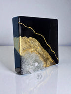 A square, black resin art piece with gold leaf accents and a geode-like crystal formation. The artwork features a glossy finish and the text "ART STUDIO 39".