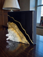 A black square art piece with gold leaf accents and a gold line design. The artwork features a cluster of clear quartz crystals on the left side. The piece is displayed on a dark wooden surface.