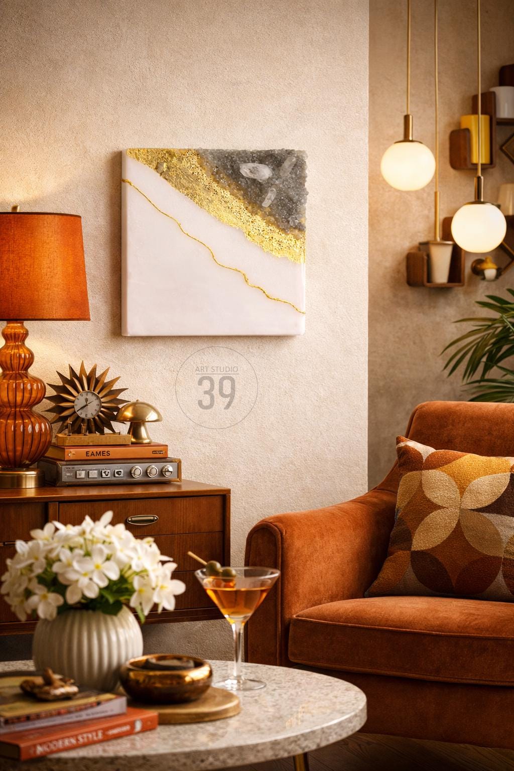 Square abstract art piece with white, gold, and gray sections. The artwork hangs on a textured wall in a room with a mid-century modern aesthetic. A lamp, clock, and cocktail are also visible.