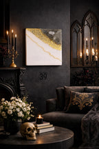 Square wall art with a white background, accented with gold leaf and a gray section. A thin gold line runs diagonally across the artwork. The image is set in a dark room with a fireplace, candles, and a sofa.