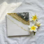 Square white resin coaster with a gold leaf and gray crystal design. A gold line accents the white surface. Three white and yellow flowers are placed on the right side of the coaster.